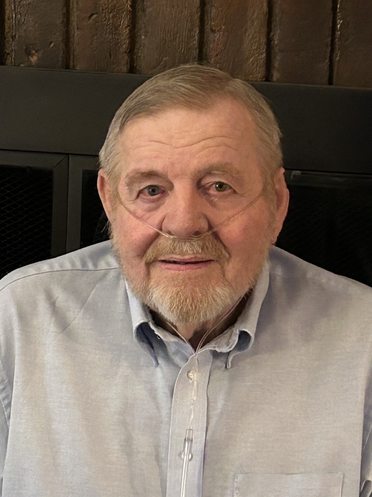 An older man with a gray beard and mustache, wearing a light blue shirt and nasal oxygen tubing, sits in front of a dark background.