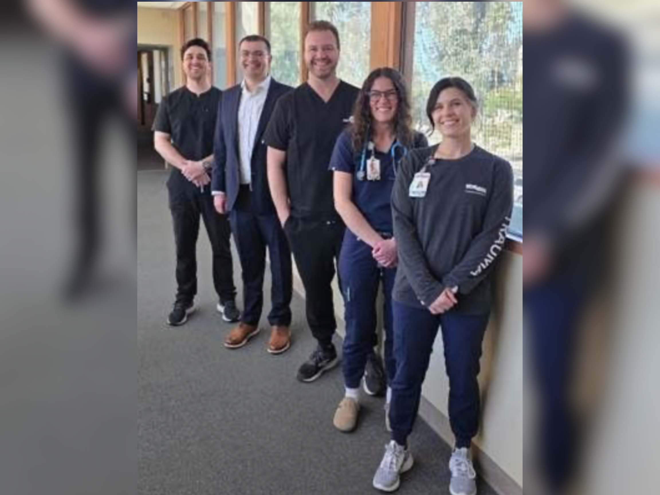 Five people, three men and two women, stand in a line indoors, wearing business and medical attire, smiling at the camera.