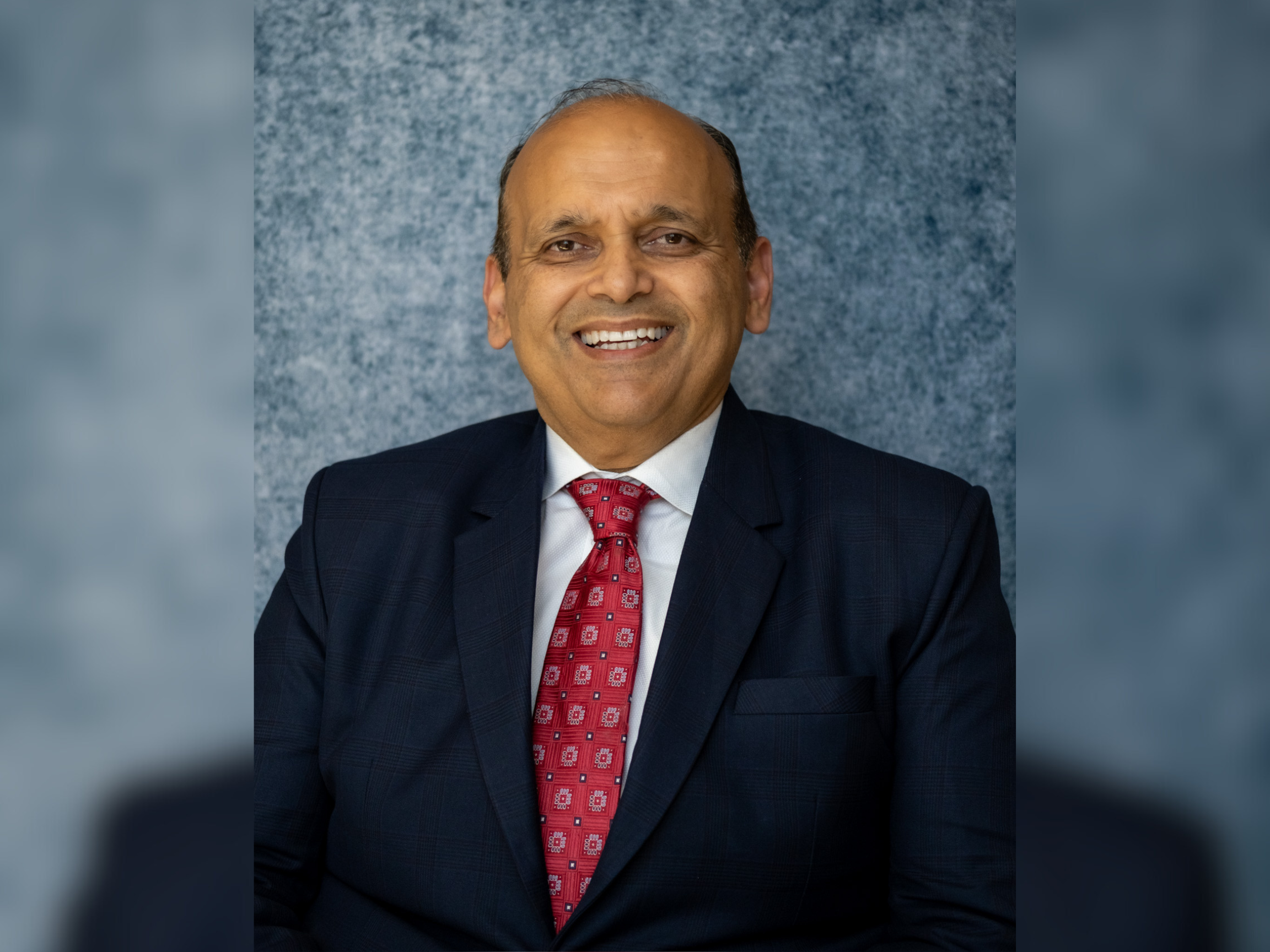 A man wearing a dark suit, white shirt, and red patterned tie smiles while sitting in front of a textured blue-gray backdrop.
