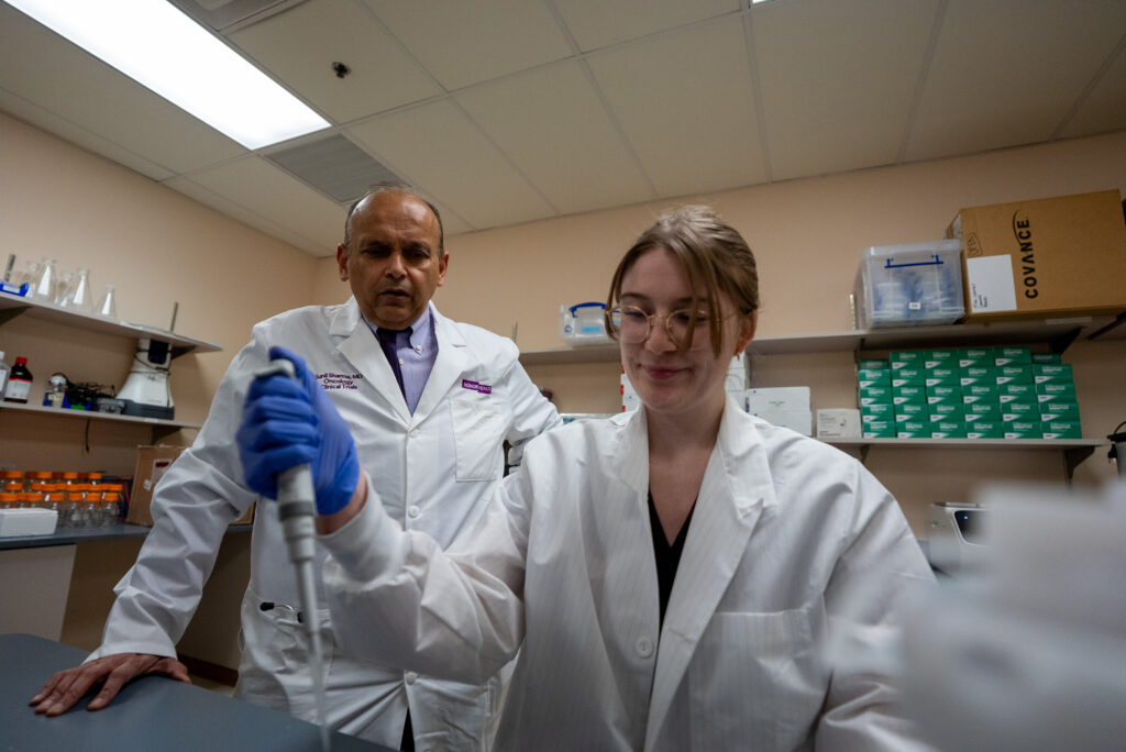 Two scientists in lab coats work in a laboratory; one operates a pipette while the other observes. Laboratory equipment and supplies are visible in the background.