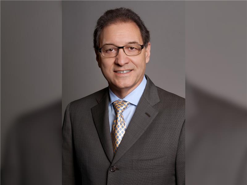 A man wearing glasses, a gray suit, blue shirt, and patterned tie poses for a professional portrait against a plain gray background.