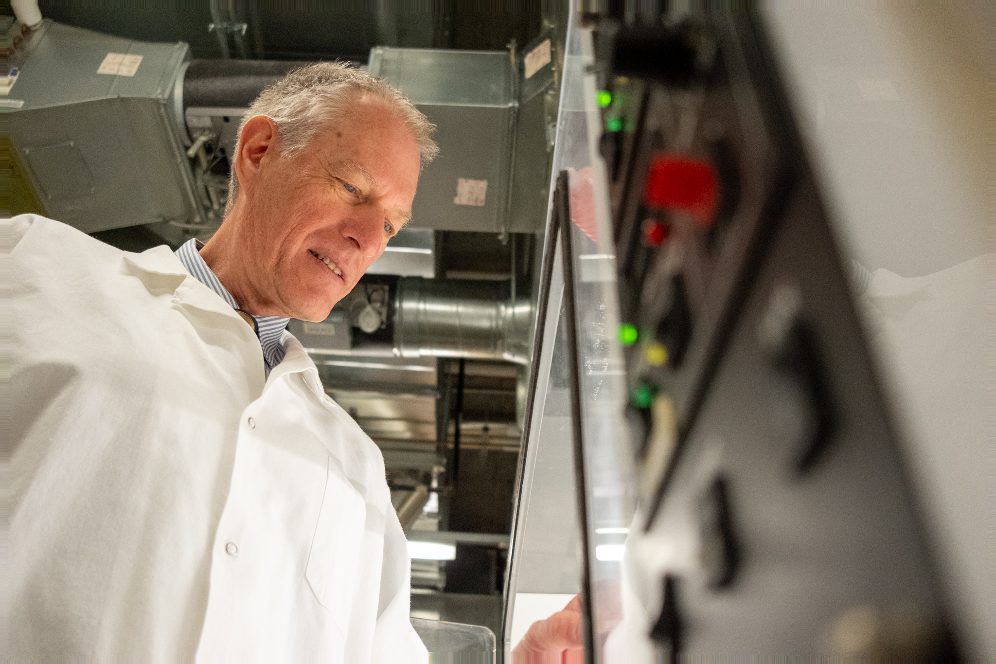A man in a white lab coat operates or inspects a control panel in an industrial or laboratory setting.