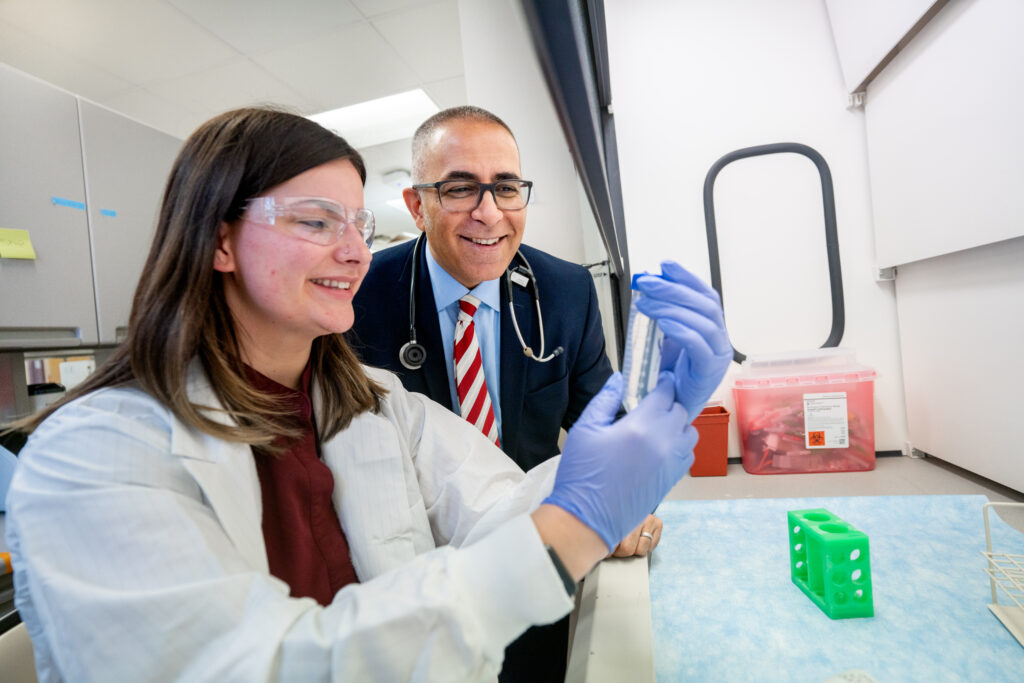 A woman with long brown hair wearing safety glasses, a white lab coat, and blue nitrile gloves holds up a test tube for examination. Beside her, a man in glasses, a navy blazer, light blue shirt, and red-striped tie smiles while observing. A stethoscope hangs around his neck. They are in a laboratory with a red biohazard sharps container and green test tube rack visible on the bench behind them.