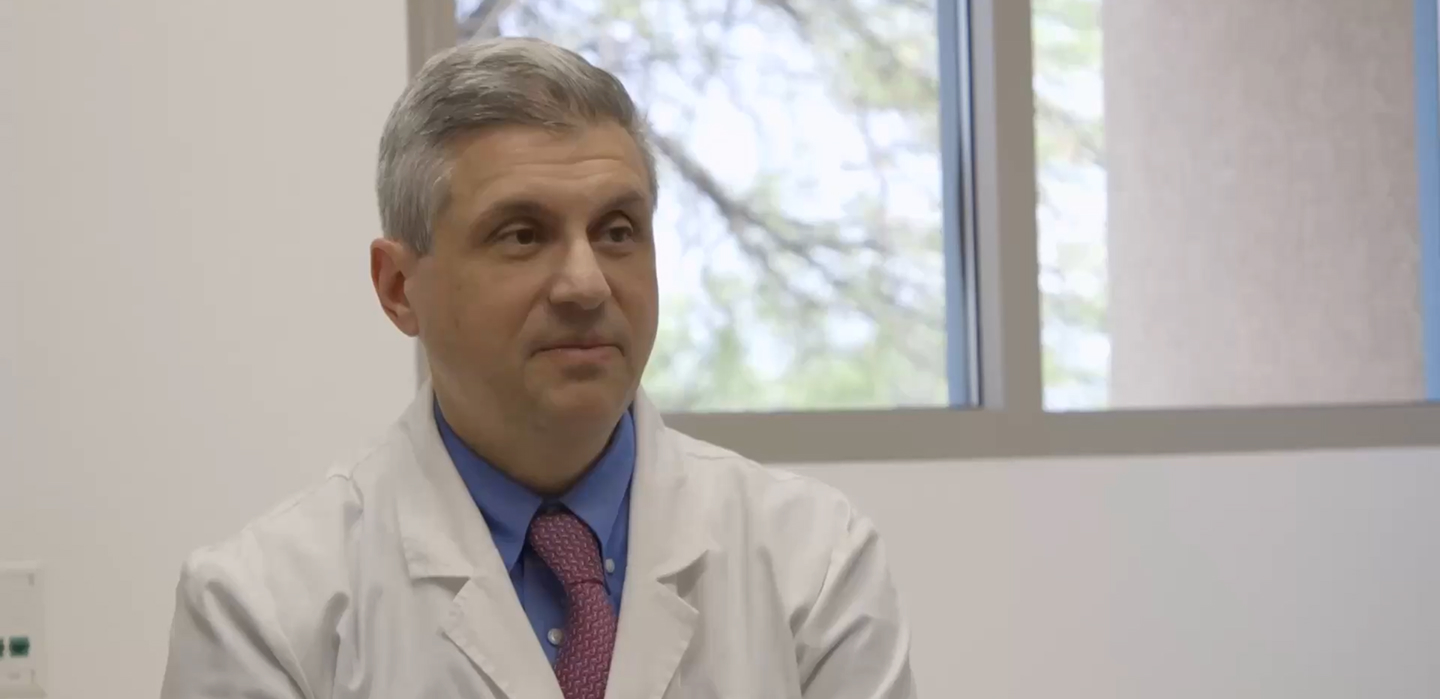A person wearing a white lab coat, blue shirt, and red tie is seated indoors with windows in the background.