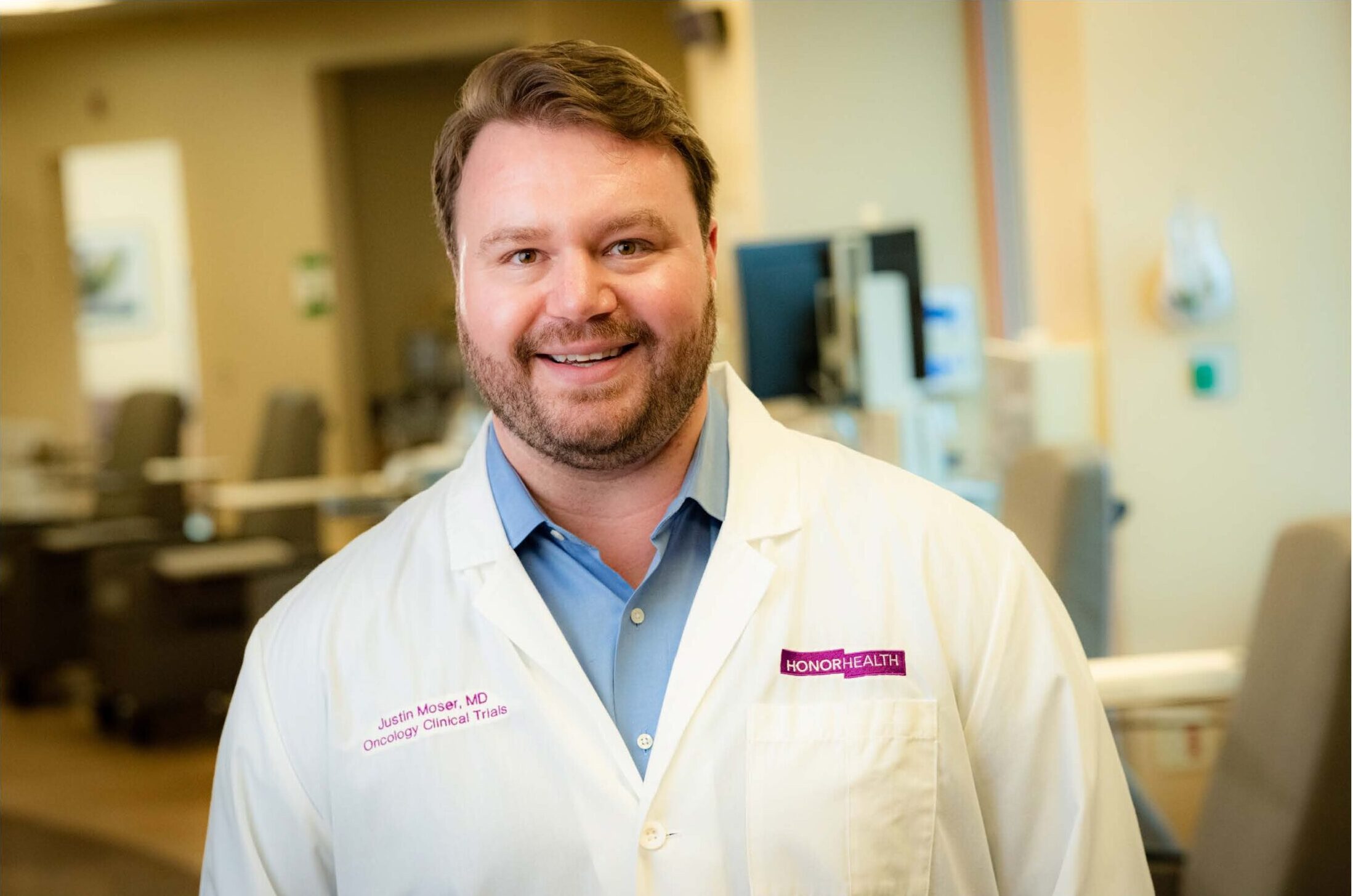 A man wearing a white lab coat with "HonorHealth" and "Oncology Clinical Trials" text stands smiling in a medical office setting.