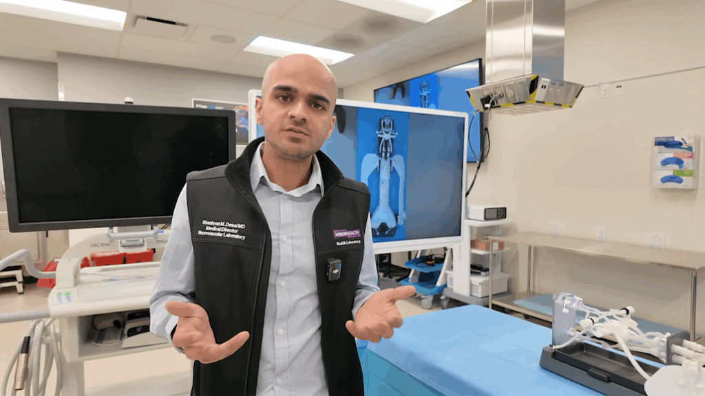 A person stands in a medical lab, wearing a black vest and gesturing with their hands. Medical equipment, monitors, and a hospital bed are visible in the background.