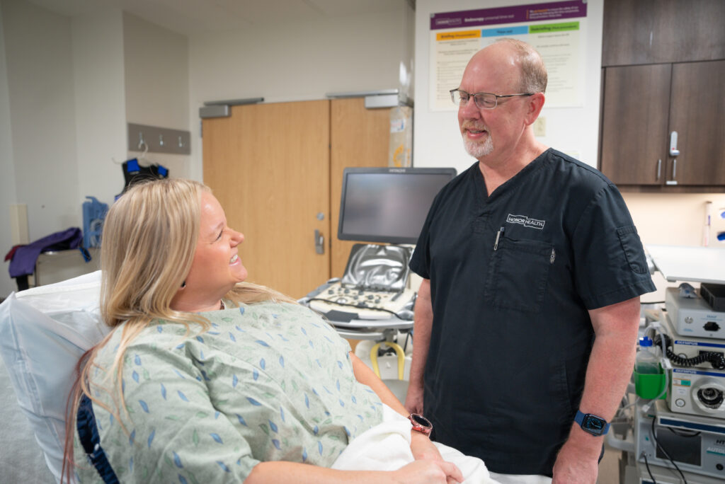 A woman in a hospital gown sits on a bed and smiles at a healthcare professional in scrubs, who is standing beside her in a medical examination room.