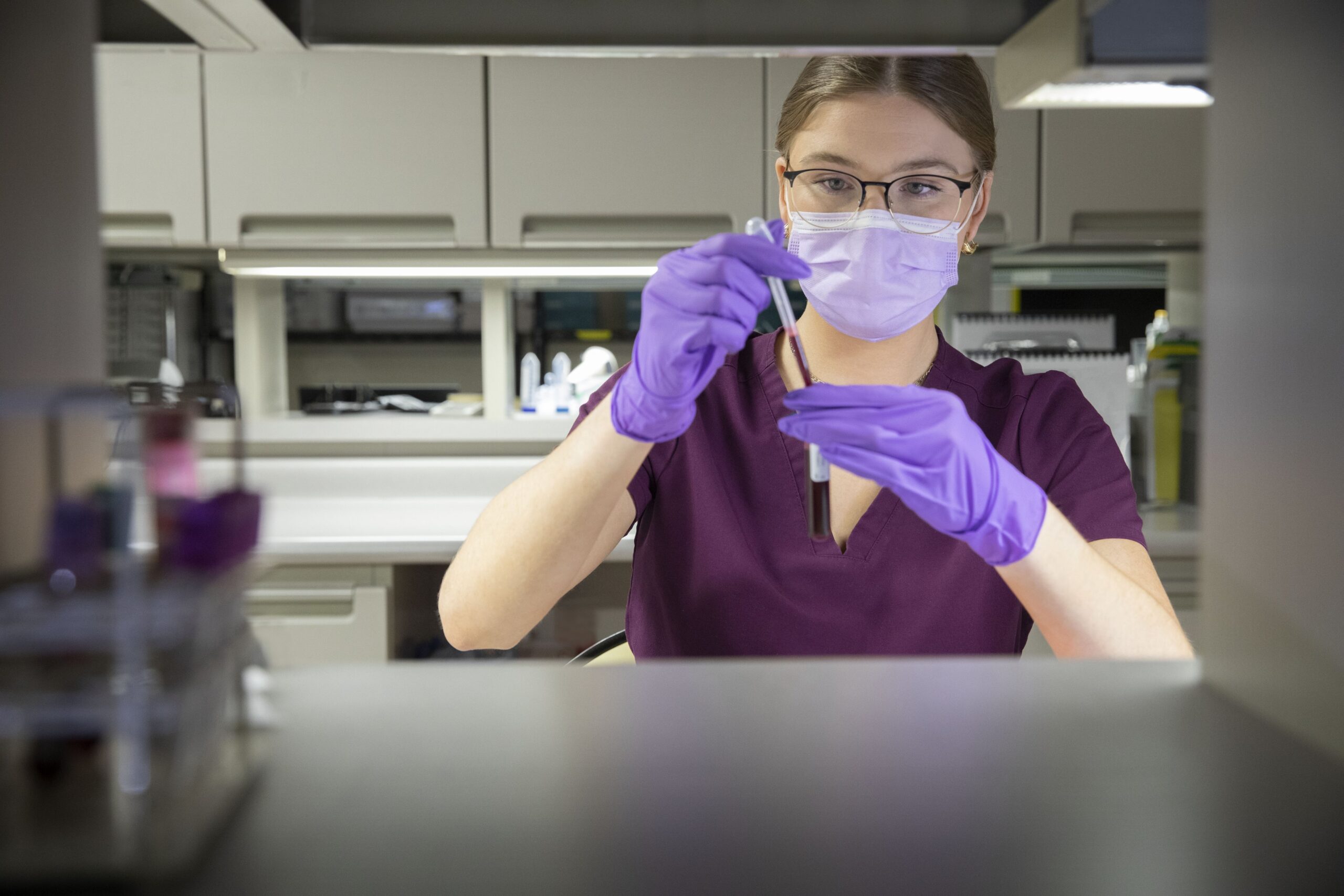 A person wearing a mask and gloves uses a pipette to transfer liquid into a test tube in a laboratory setting.