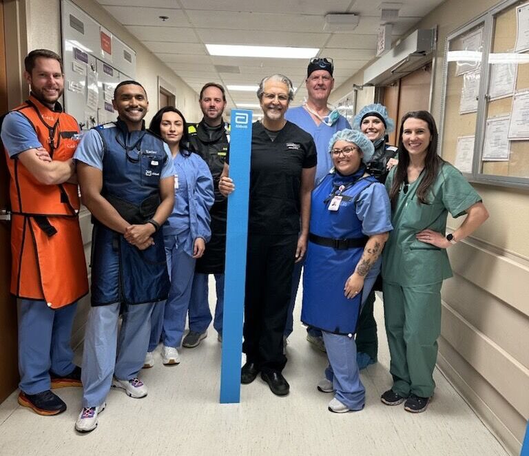 A group of nine medical professionals in scrubs and uniforms stand together and smile for a photo in a hospital hallway.