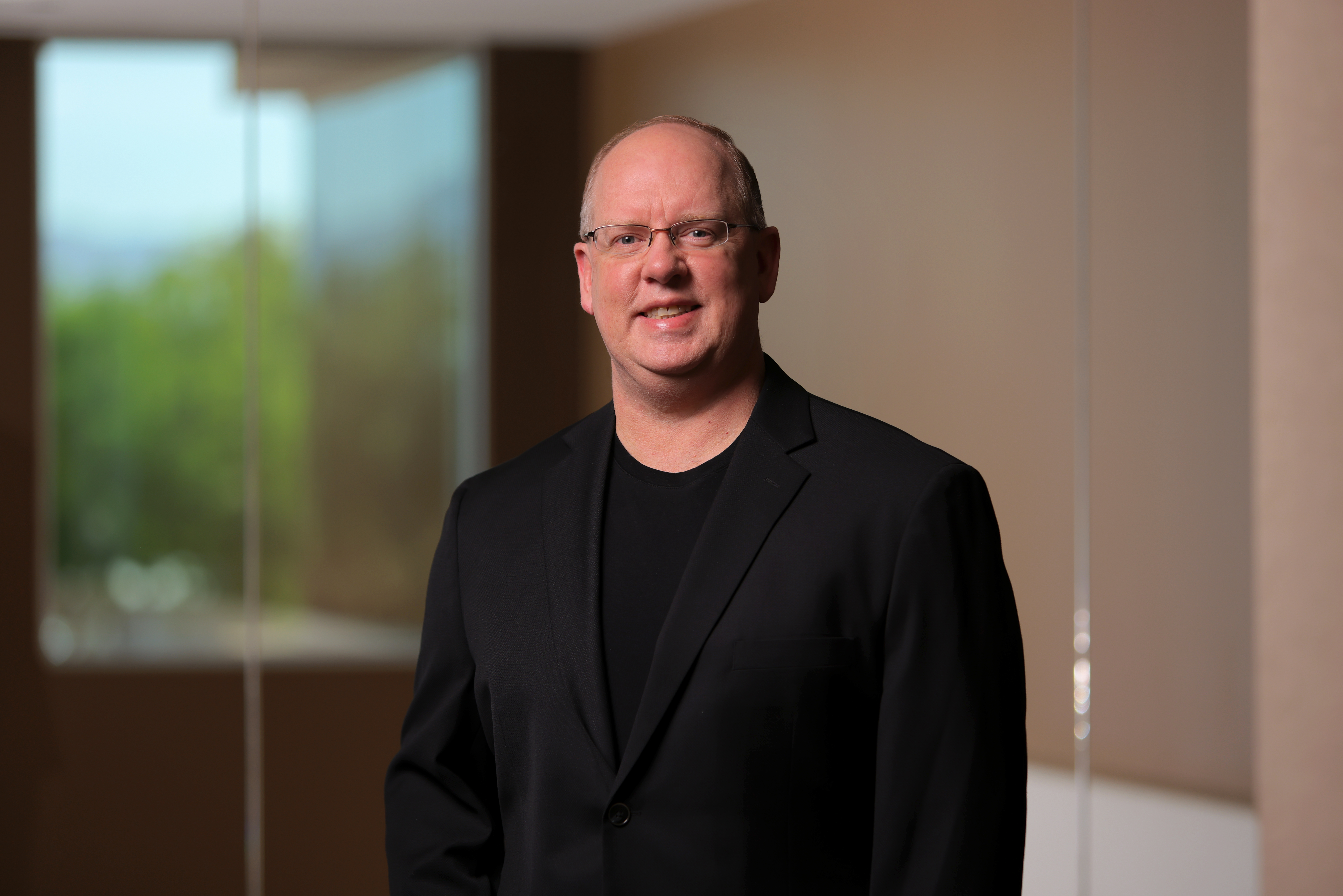 A man wearing glasses and a black suit jacket over a black shirt stands indoors in front of a blurred background with windows.