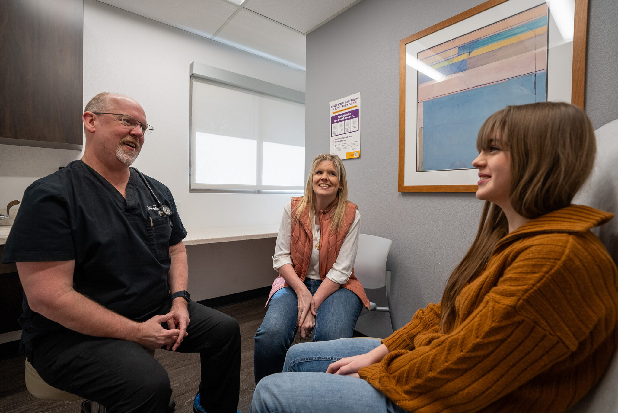 A doctor talks with a woman and a teenage girl in a medical exam room. The woman sits in a chair, the girl sits on the exam table, and the doctor is facing them.