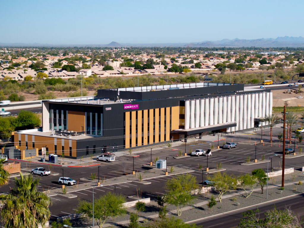 A modern two-story medical building labeled “HonorHealth” sits in a large parking lot with several cars, surrounded by trees and suburban homes in the background.