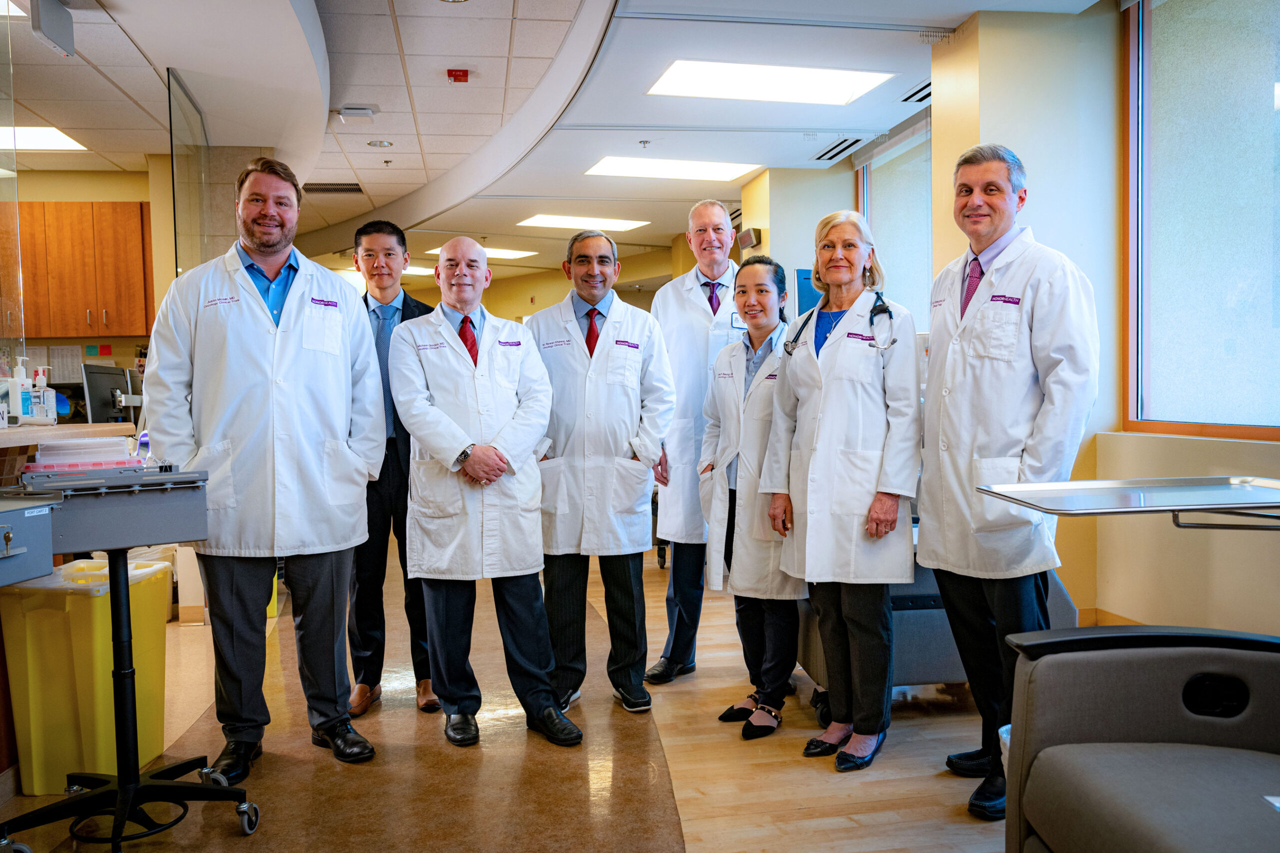 A group of eight medical professionals in white lab coats stand together in a hospital hallway, posing for a photo.
