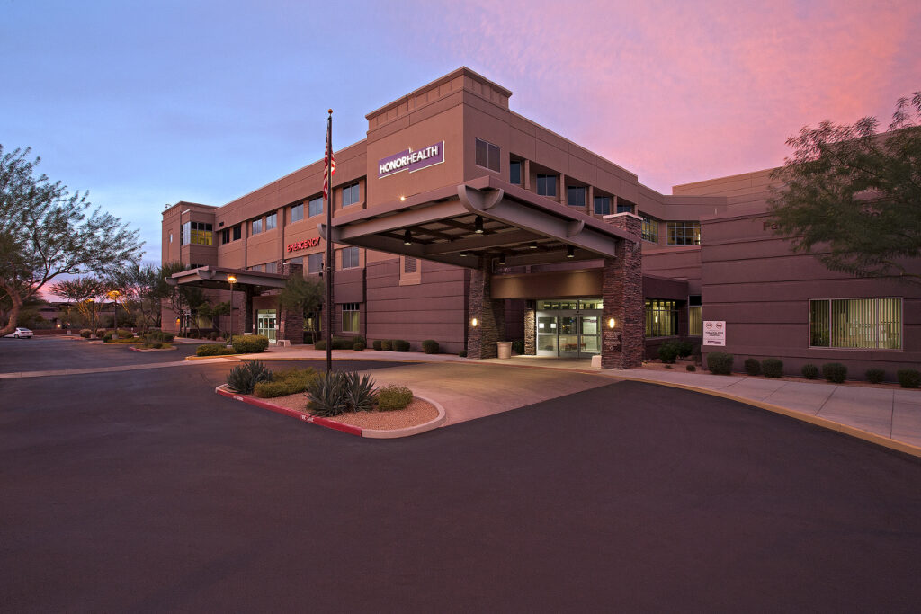 A modern two-story medical center building with an entrance canopy, surrounded by landscaped grounds and an American flag at sunset.