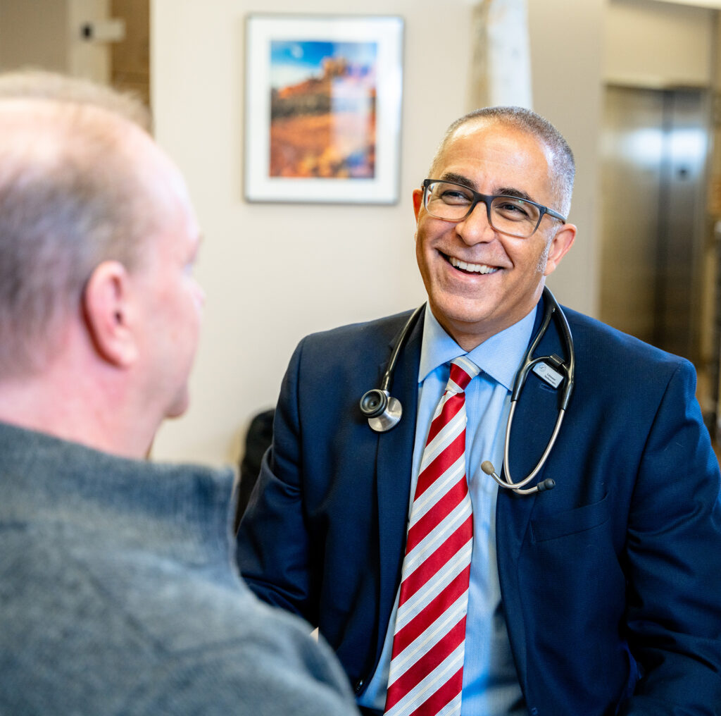 A doctor wearing a suit, striped tie, and stethoscope smiles while talking to a patient in an office setting.