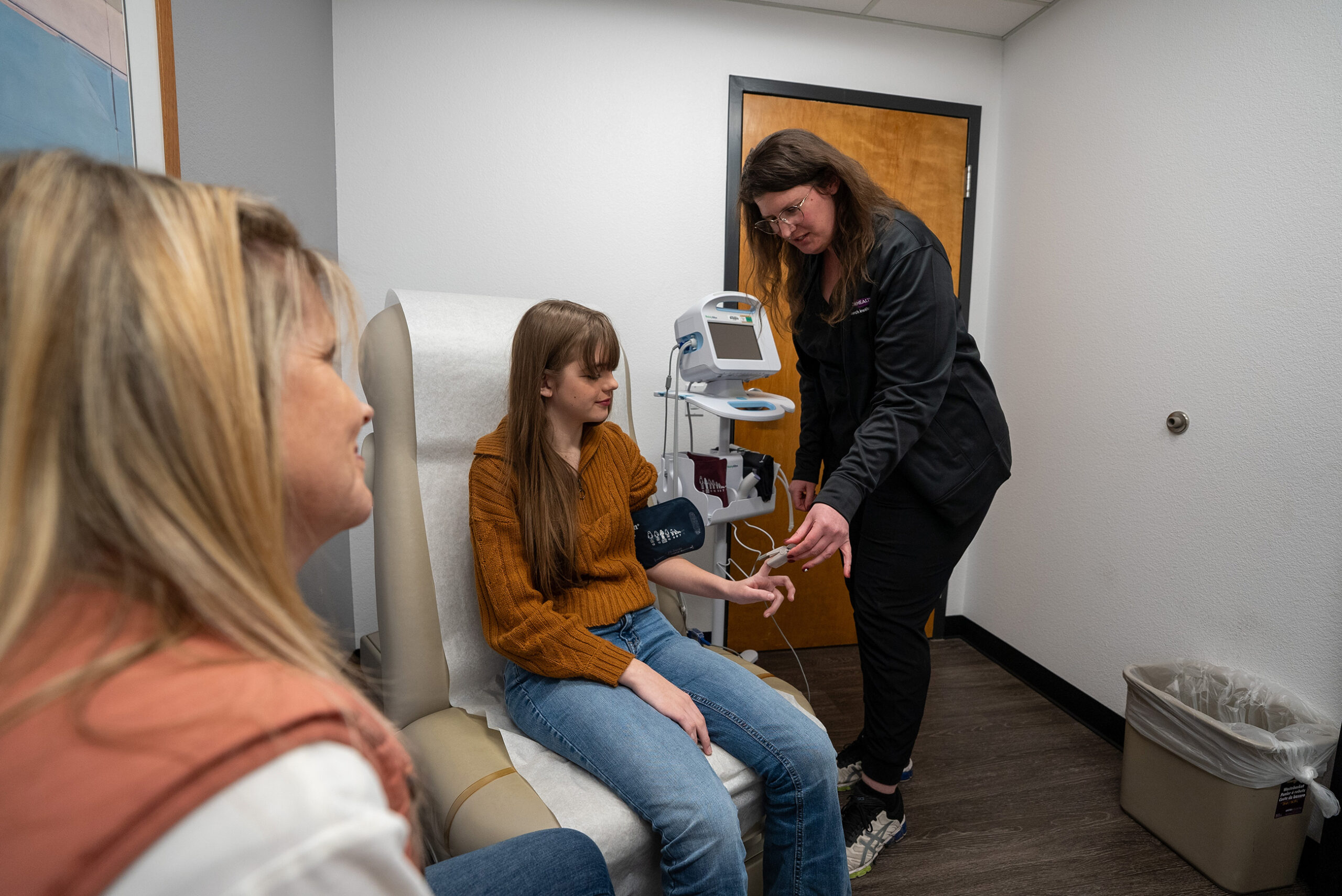 A healthcare worker checks a young woman's blood pressure in a medical exam room while another woman sits nearby.