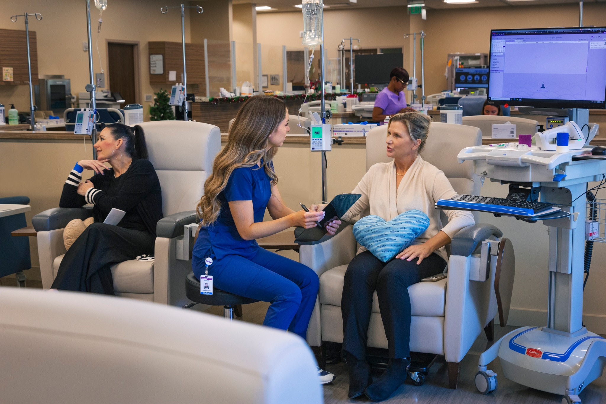 A healthcare worker in scrubs speaks with a seated patient holding a heart-shaped pillow in a medical treatment area, with other patients and monitors visible.