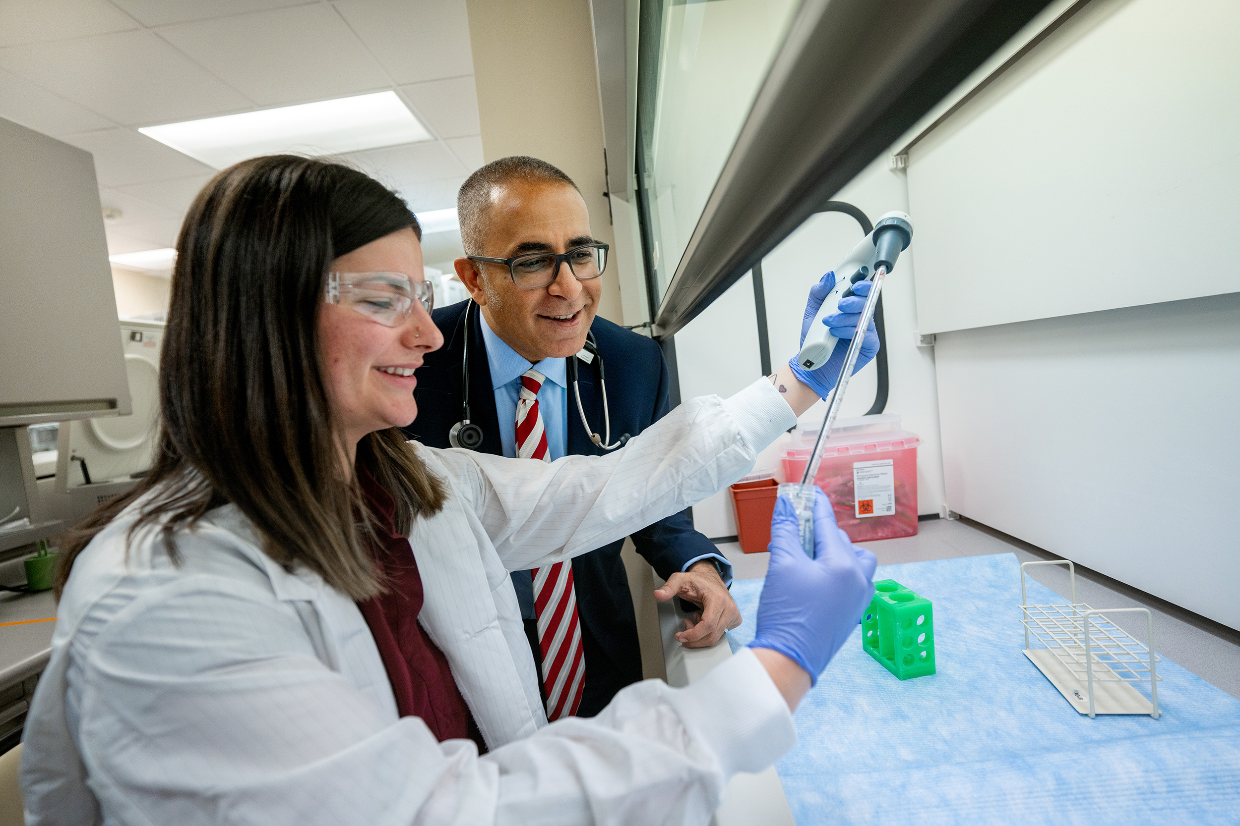 A woman in a lab coat uses a pipette while a man in a suit with a stethoscope observes in a laboratory setting.