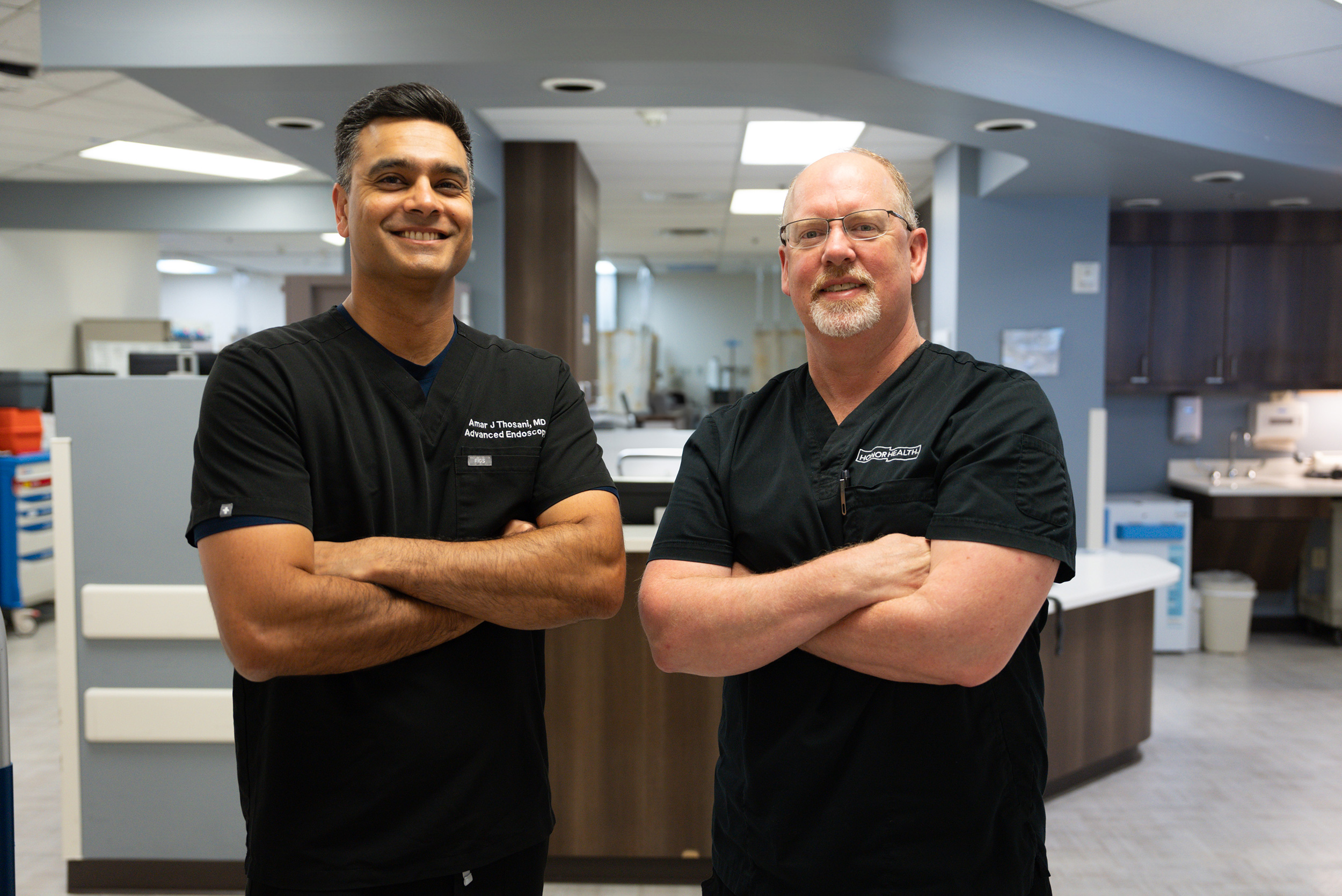Two healthcare professionals in black scrubs stand with arms crossed and smile in a modern medical facility.
