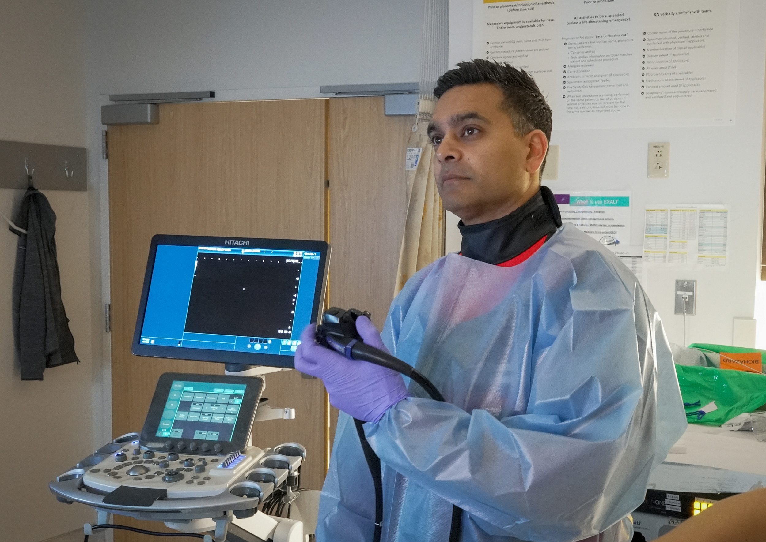 A healthcare professional wearing protective gear holds an endoscope next to an ultrasound machine in a medical examination room.