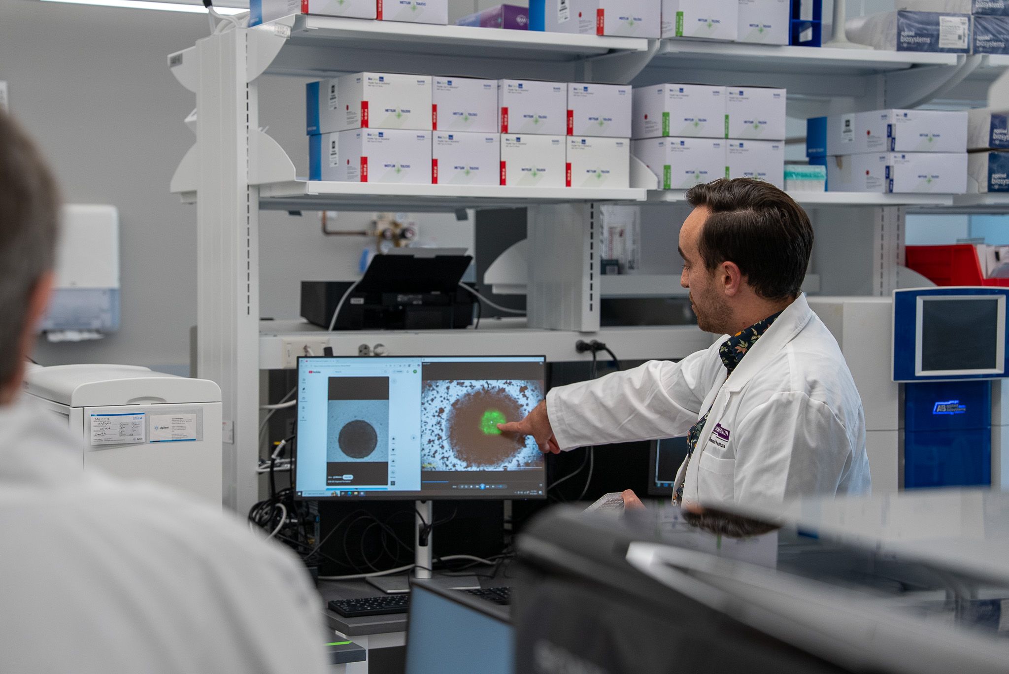 A scientist in a lab coat points to results displayed on a computer monitor, showing a circular pattern, while another person observes in a laboratory setting.