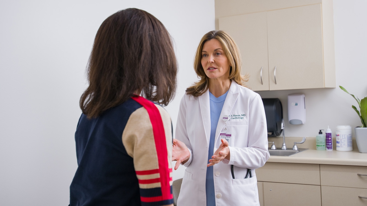 A doctor in a white coat speaks with a patient in an exam room, gesturing with her hands. Medical supplies are visible on the countertop in the background.