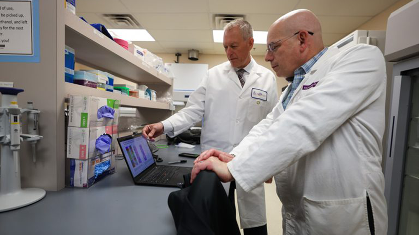 Two scientists in lab coats examine data displayed on a laptop screen in a laboratory setting.