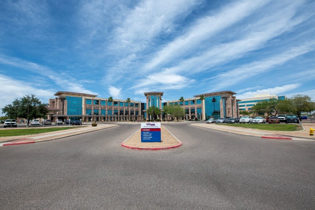 A large modern office building with blue-tinted windows and palm trees, seen from a driveway entrance with a directional sign in the center foreground.