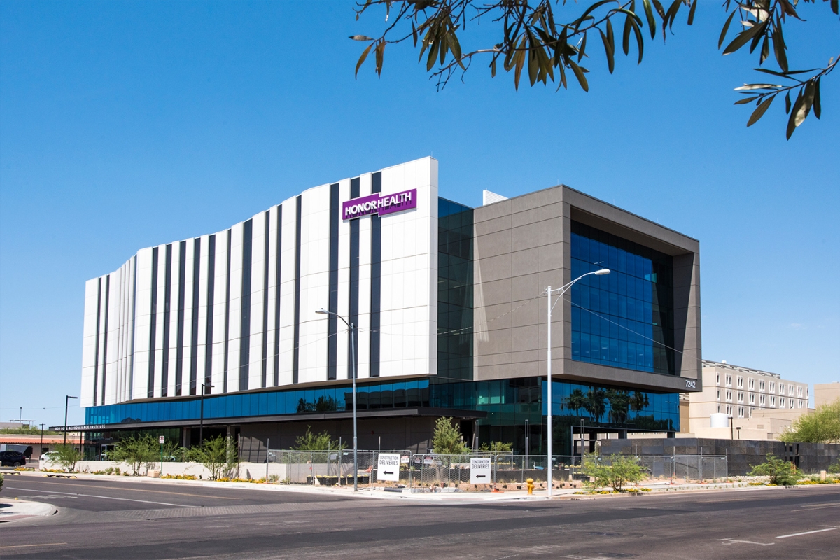 A modern, multi-story building with a "HonorHealth" sign, featuring glass and geometric architecture, stands by a street under a clear blue sky.