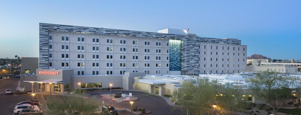 A modern multi-story hospital building with an illuminated emergency entrance sign at dusk, surrounded by trees and a parking area.