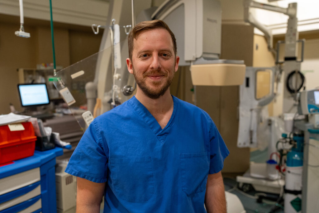 A person wearing blue medical scrubs stands in a hospital or clinical setting, surrounded by medical equipment and supplies.
