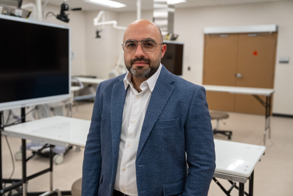 A man in a blue blazer and white shirt stands in a modern, well-lit laboratory or medical classroom with tables and equipment in the background.