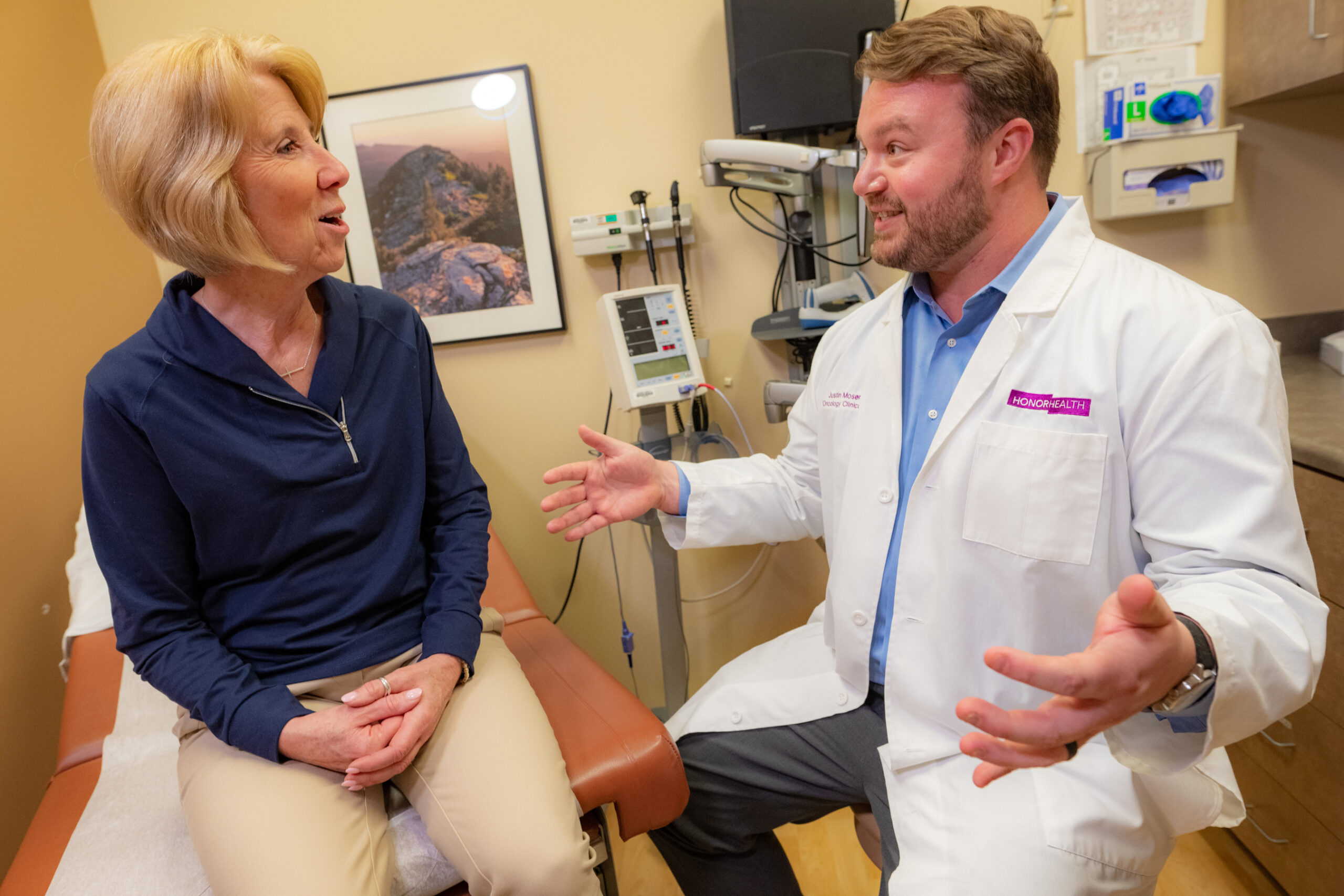 A doctor in a white coat gestures while talking to a female patient seated on an exam table in a medical office.