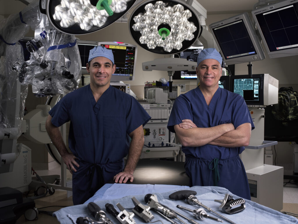 Two surgeons in blue scrubs stand in an operating room with surgical tools laid out on a table and medical equipment in the background.