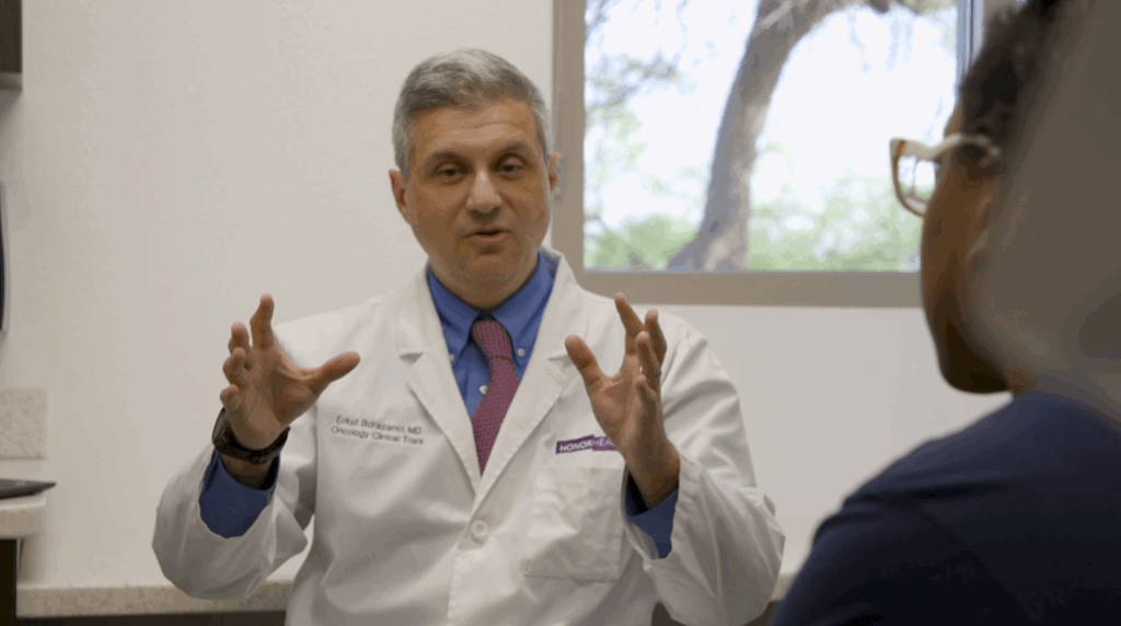 A doctor in a white coat gestures with both hands while speaking to a seated patient in a medical office with a window in the background.