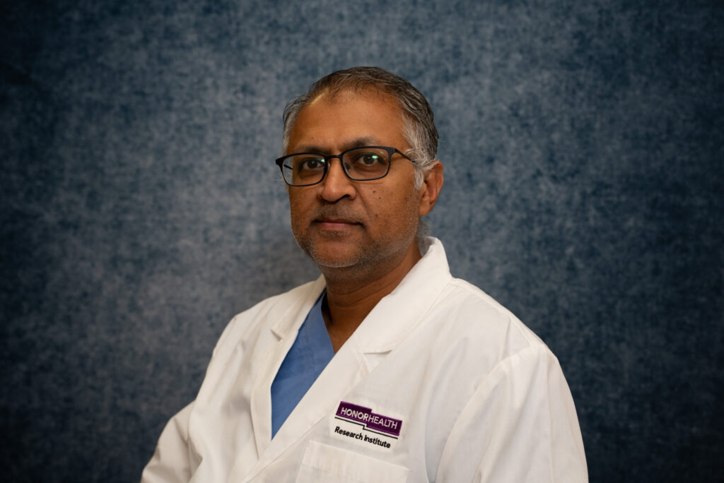 A man wearing glasses and a white lab coat stands in front of a textured blue background. The lab coat displays the HonorHealth logo and a name tag.