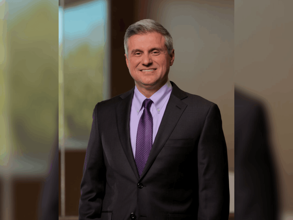 A man in a dark suit and purple tie stands indoors, smiling, with a blurred background featuring a window and greenery outside.