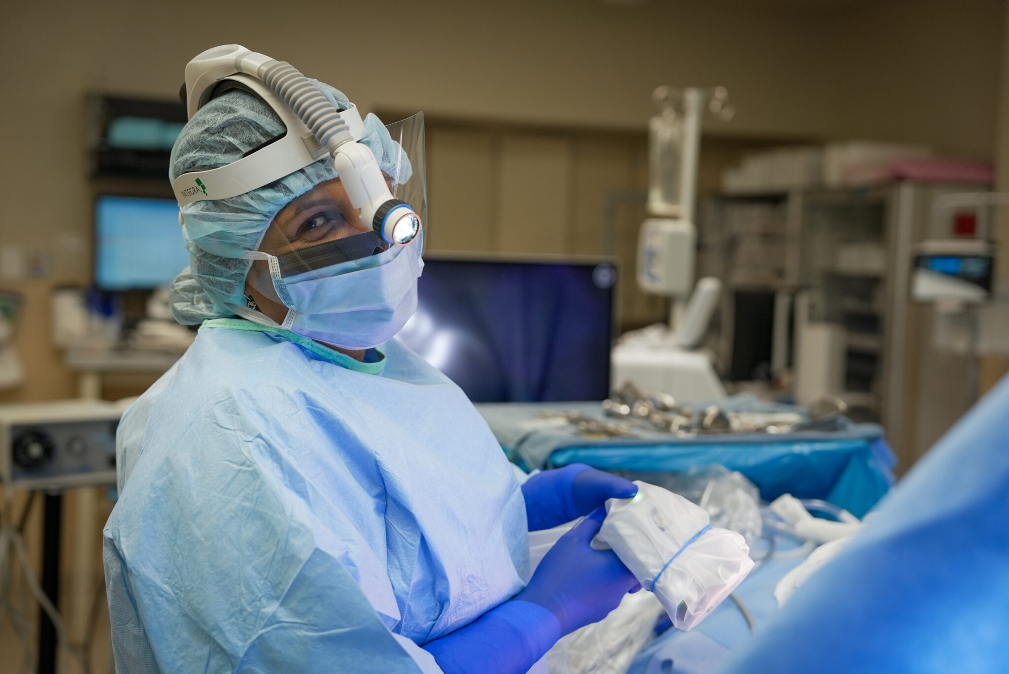 A medical professional in surgical attire and protective gear is holding a device in an operating room, surrounded by medical equipment.