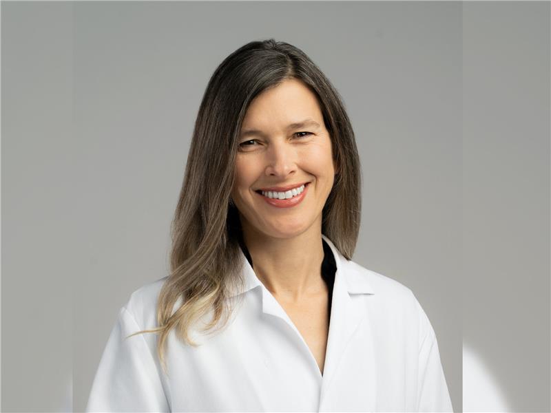 A woman with long brown hair wearing a white lab coat smiles at the camera against a plain light gray background.