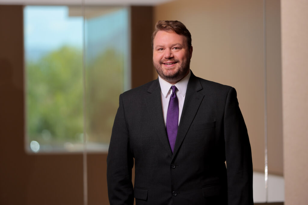 A man in a dark suit and purple tie stands indoors in front of a blurred background with large windows, as he prepares to present breakthrough research on synovial sarcoma immune-cell therapy.